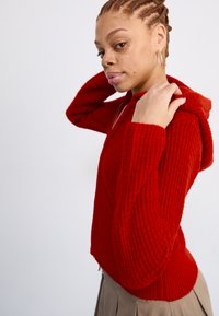 Rode gebreide hoodie met ribbelstructuur, lange mouwen en een voorkantzak. Voorzien van een capuchon met trekkoord en een iets verkorte ontwerp.