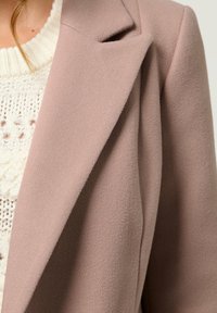Lichtroze wollen blend jas met een geknipt kraag, getailleerde pasvorm en een zachte, gestructureerde oppervlakte, gelaagd over een crème gebreide trui.