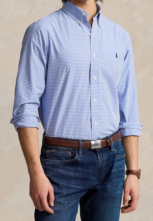 Homme portant une chemise à petits carreaux bleu et blanc à boutons, une ceinture en cuir marron, un jean bleu et une montre-bracelet marron.