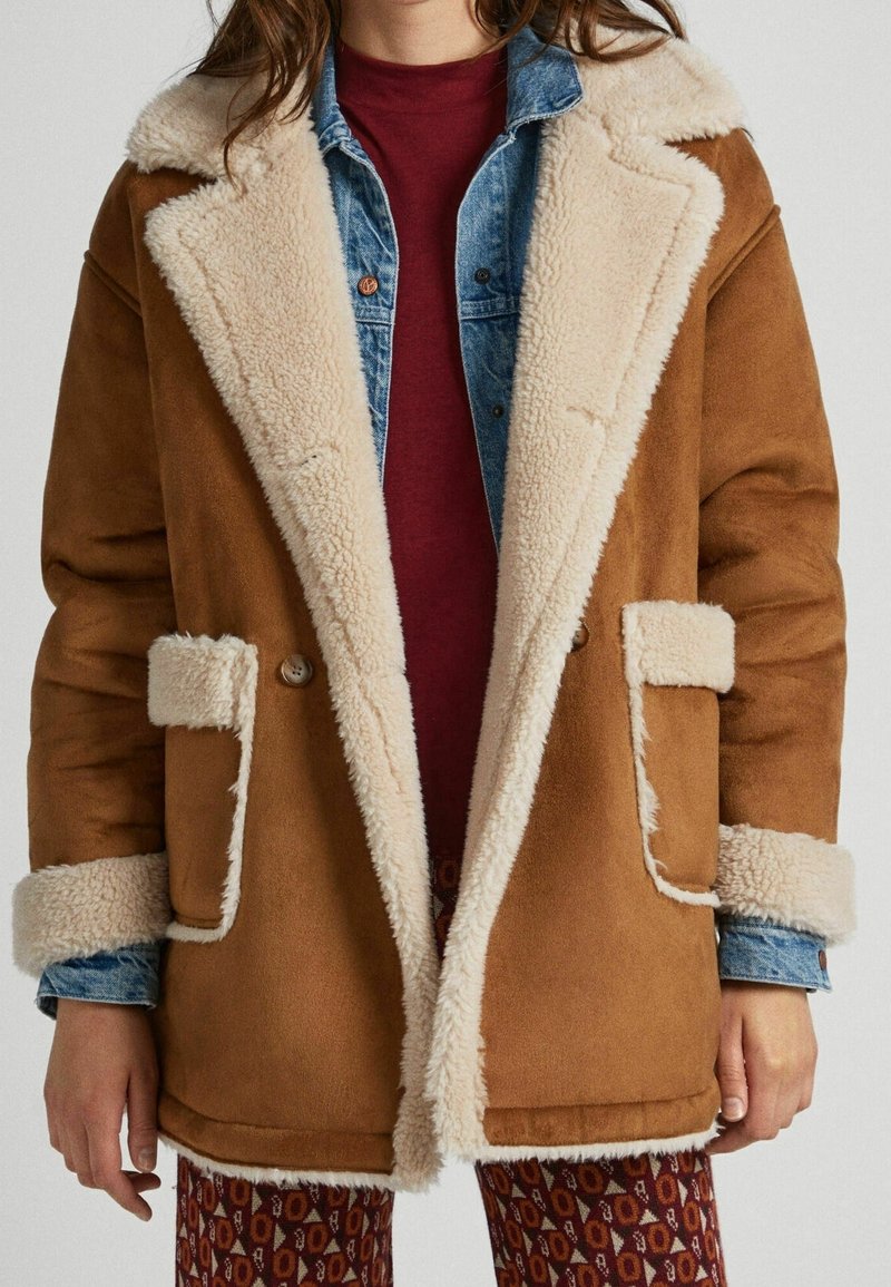 Brun shearling-kappa med en fluffig krämfärgad krage och manschetter, dubbelknäppt framsida och två sidofickor, lager på lager över en denimskjorta.