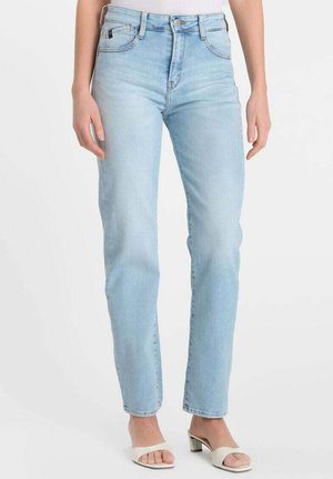 Slim-fit lichtblauwe spijkerbroek gedragen door een staand persoon met een lichte huid, gecombineerd met witte open-teen sandalen met blokhak.