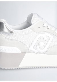 Sneaker bianca con tomaia in tessuto testurizzato, dettagli in suede e un particolare sul tallone argentato. Presenta un logo bianco pronunciato e una suola spessa.