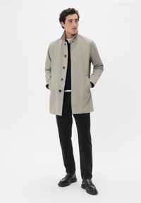 Chaqueta exterior de un beige claro con collar alzado, cierre de botones negros y bolsillos laterales, combinada con pantalones y zapatos negros.