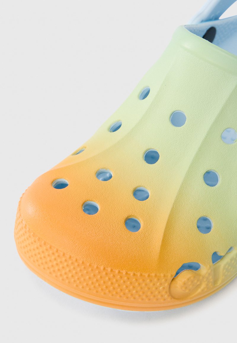 Zueco con degradado de verde a naranja, con orificios de ventilación, superficie texturizada y un material suave y duradero. Presenta una suela estampada para mayor tracción.