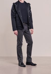 Svart vattentät jacka med huva, lager-på-lager över en mörk tröja, parat med grå byxor och svarta läderstövlar. Har dragkedja och knappar.