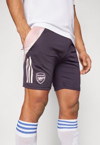 Pantalones cortos deportivos de color gris oscuro con paneles laterales rosas, que presentan rayas blancas y un logo del Arsenal en el muslo izquierdo. Tela suave y ligera.