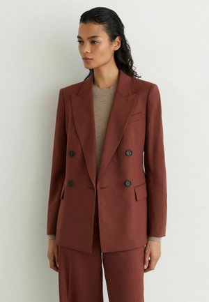 Frau mit dunklem Haar trägt einen rostfarbenen, zweireihigen Blazer und passende Hose über einem beigen Oberteil und steht vor einer weißen Wand.