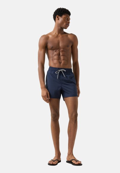 Atletisk mand iført marineblå badeshorts med snor og sorte klipklapper, stående med hovedet vendt til siden, på en hvid baggrund.