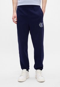 Tengerész sötétkék melegítőnadrág, pamut keverék anyagból készült, rugalmas derékkal, lekerekített bokákkal, és fehér logónyomattal a combon.