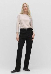 Helles Rosa Langarmshirt kombiniert mit schwarzen maßgeschneiderten Hosen. Das Outfit verfügt über einen schwarzen Gürtel mit einem silbernen Emblem und flachen schwarzen Schuhen.