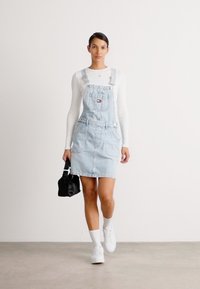 Lys blå denim dungaree-kjole med justerbare stropper, frontlomme, sidelommer og en tettsittende design, kombinert med en svart veske og hvite sneakers.