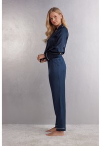 Navy blauwe satijnen pyjama set met lange mouwen, elastische tailleband en witte piping langs de kraag en manchetten; model staat blootsvoets op het tapijt.