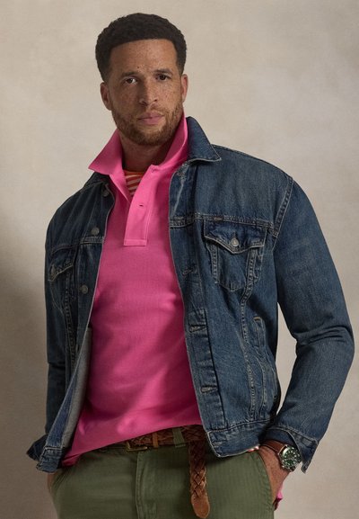 Τζιν μπουφάν πάνω από ροζ πόλο με ριγέ εσωτερικό γιακά, πράσινο παντελόνι και πλεκτή ζώνη, με ρολόι στο χέρι.