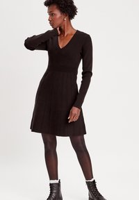 Schwarzes geripptes Kleid mit langen Ärmeln, V-Ausschnitt, tailliertem Oberteil und ausgestelltem, plissiertem Rock. Kombiniert mit schwarzen Schnürstiefeln und weißen Socken.