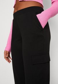 Černé kalhoty s hladkým povrchem, vybavené boční kapsou na náklad. Růžový žebrovaný top s dlouhým rukávem, částečně viditelný na horní straně.