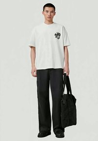 T-shirt branca de manga curta com gráfico preto, combinada com calças cargo soltas pretas e uma tote bag preta, com alças resistentes e fechos de correr.