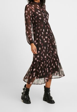 Kobieta ubrana w czarną przezroczystą sukienkę midi z różowymi abstrakcyjnymi plamami i długimi rękawami, połączoną z czarnymi skórzanymi botkami na kostkę z klamrą.