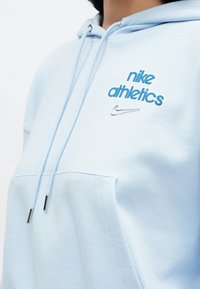Ανοιχτό μπλε φούτερ με κουκούλα, με κεντημένο το «nike athletics» και το λογότυπο της Nike σε μπλε. Μαλακή υφή και λεπτομέρεια με τσέπη μπροστά.