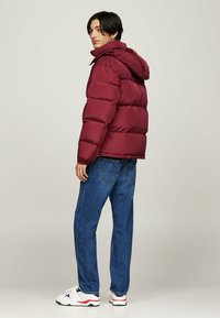 Jachetă puffer bordo cu glugă, textură strălucitoare și matlasată, asociată cu blugi albaștri și adidași albi cu accente roșii, văzută din spate.