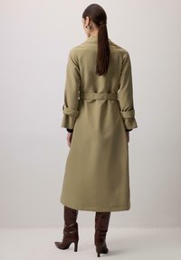 Olivnozeleni trenčkot z obložitim pasom, dolgimi rokavi in zavihanimi manšetami, ter srednje dolžine. Gladek, teksturiran material.