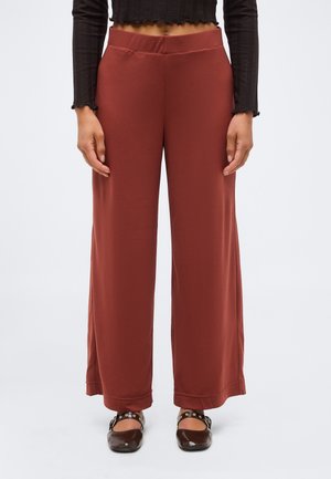 Frau trägt rostfarbene, weit geschnittene Hosen kombiniert mit einem schwarzen, langärmeligen, kurz geschnittenen Oberteil und dunkelbraunen flachen Schuhen mit Riemen.