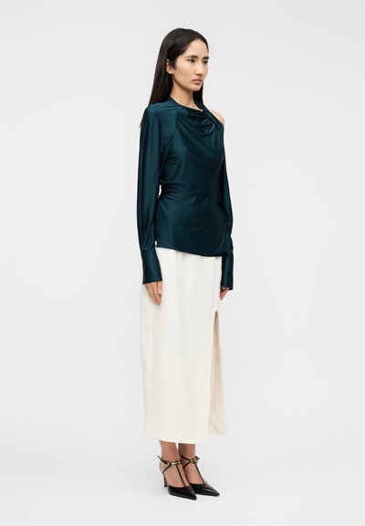 Γυναίκα που φοράει σκούρο τιρκουάζ ασύμμετρο τοπ με μακρύ μανίκι και έναν ακάλυπτο ώμο, λευκή μίντι φούστα με σκίσιμο και μαύρες μυτερές γόβες, όρθια πλάγια.