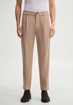Homme portant un pantalon plissé beige, des chaussures noires et une chemise blanche rentrée, debout devant un fond uni.