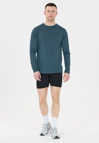 Sportliches langärmliges Shirt in dunkeltürkis, aus strukturiertem Stoff. Kombiniert mit schwarzen Shorts und hellen Sneakers mit weißen Socken.