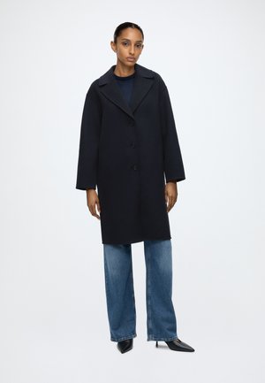 Donna in piedi che indossa un cappotto nero abbottonato, jeans blu larghi e tacchi neri appuntiti su uno sfondo bianco uniforme.