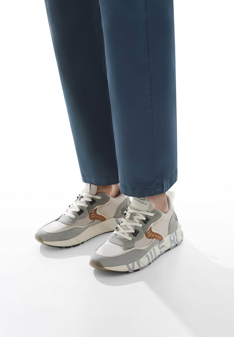 Weiße und graue Sneakers mit tanfarbenen Akzenten, strukturierter Textiloberfläche und einem gemusterten Profil. Kombination mit dunkelblauen Hosen, der Fokus liegt auf den Details des Schuhwerks.