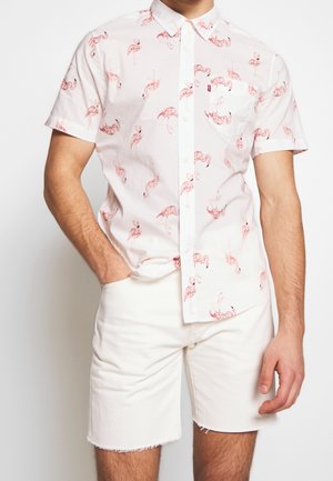Chemise à manches courtes avec boutons en tissu blanc ornée d'un motif de flamants roses. Comprend une poche poitrine et un col classique. Portée avec un short blanc.