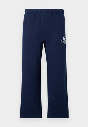 Deux personnes portant des t-shirts blancs Lacoste, des pantalons de survêtement Lacoste bleu marine et des baskets blanches, debout devant un fond gris uni.