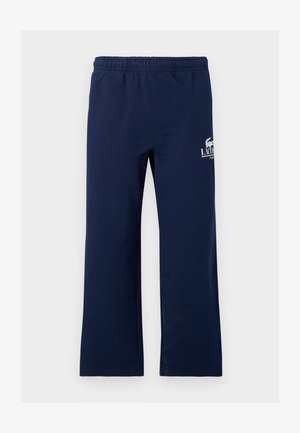 Deux personnes portant des t-shirts blancs Lacoste, des pantalons de survêtement Lacoste bleu marine et des baskets blanches, debout devant un fond gris uni.