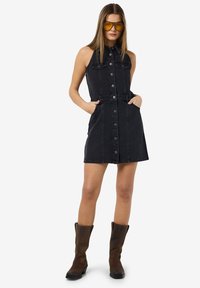 Zwart denim mouwloos jurkje met knoopsluiting aan de voorkant, zijzakken en een getailleerde taille. Gecombineerd met bruine kniehoge laarzen.