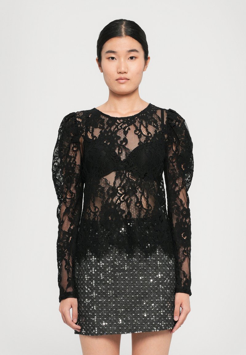 Femme portant un haut en dentelle noire à manches longues avec des épaules bouffantes et une mini-jupe noire à sequins, se tenant devant un fond uni.