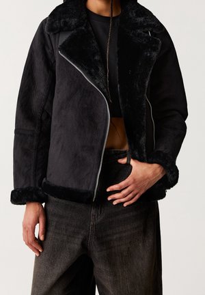 Personne portant un blouson en fausse fourrure noire avec une fermeture éclair diagonale sur un haut noir et un jean foncé, la main reposant sur la ceinture.