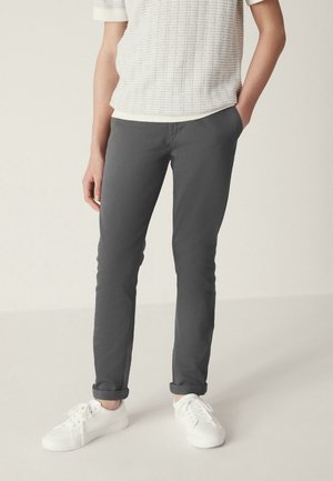 Person wearing gray slim-fit pants with cuffs, a white textured short-sleeve top, and white sneakers, standing with one hand in pocket.