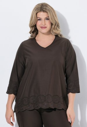 Mujer con cabello rubio ondulado que lleva una blusa marrón oscuro con cuello en V, dobladillo festoneado bordado y mangas 3/4 sobre un fondo claro y liso.