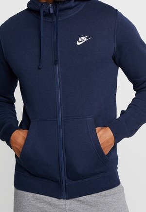 Man draagt een donkerblauwe Nike zip-up hoodie met voorzakken en capuchon, handen in de zakken, gecombineerd met grijze joggingbroek.