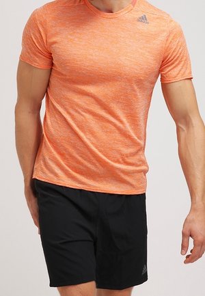 Mand iført orange Adidas kortærmet atletiktrøje og sorte Adidas shorts, står mod en ensfarvet lys baggrund.