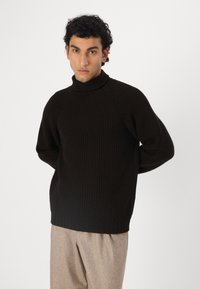 Donkerbruine geribbelde gebreide trui met hoge kraag en raglanmouwen, gecombineerd met lichtbruine broek. Zachte textuur en een losse pasvorm.