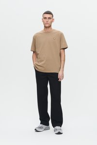 Junger Mann steht mit den Händen in den Taschen, trägt ein beiges T-Shirt, schwarze Hosen und graue Sneaker vor einem schlichten weißen Hintergrund.