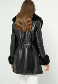 Cappotto in pelle nera con vita rifinita da una cintura, colletto e polsini in pelliccia sintetica, e una texture liscia. Presenta un taglio di lunghezza media e un design su misura.