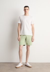 Jeune homme debout portant un t-shirt blanc uni, un short vert clair et des baskets blanches sur un fond neutre.