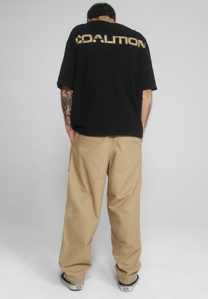Mężczyzna stojący tyłem, ubrany w czarną koszulkę z napisem "COALITION", beżowe spodnie i czarne trampki, na tle jednolitego jasnego tła.