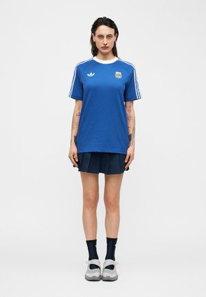 Frau steht frontal, trägt ein blaues Adidas Argentina Fußballtrikot, einen marineblauen Faltenrock, schwarze Socken und weiße Turnschuhe.