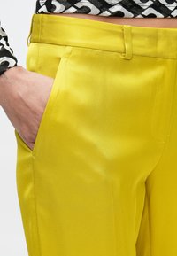 Pantalones de satén amarillos con un corte ajustado, bolsillos laterales y una textura suave, con cintura alta y sin cierres visibles.