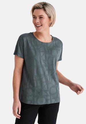 Lächende Frau, die ein kurzärmeliges, dunkelgrünes Oberteil mit Muster und schwarze Hose trägt, steht vor einem schlichten hellen Hintergrund.