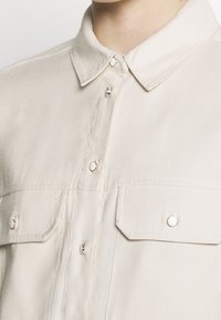Camisa beige de botones fabricada en tejido suave, con un cuello clásico, dos bolsillos en el pecho y botones de presión plateados.