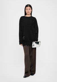 Black oversized sweater with textured accents, paired with wide brown trousers and a reflective silver clutch. Flat black shoes complete the look.